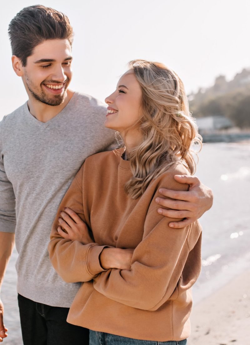 Pleasant young man embracing girlfriend on nature background. Outdoor photo of pleased blonde girl posing at sea with husband.